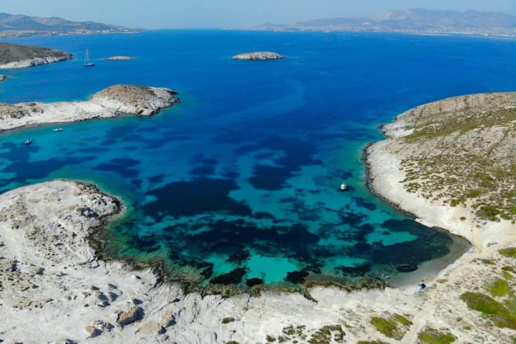 Aerial view of Faneromeni Beach on Antiparos, Greece, showcasing a secluded bay with clear turquoise and deep blue waters surrounded by rocky, white shoreline. Small boats are anchored nearby, highlighting the remote and serene beauty of this coastal area
