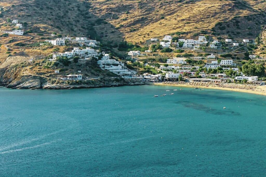 Scenic view of Ios Island, Greece, with whitewashed buildings scattered across the hillside overlooking a calm, turquoise bay. The sandy beach, dotted with small boats, lies nestled between rugged hills, showcasing the tranquil charm of this Greek island coastline.