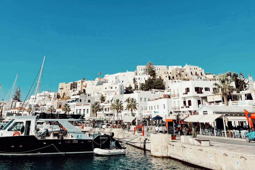 Harbor view of Naxos Island, Greece, featuring traditional whitewashed buildings stacked along the hillside under a clear blue sky. Boats are docked along the waterfront, with cafes and shops lining the promenade, capturing the charm of this Greek island town.