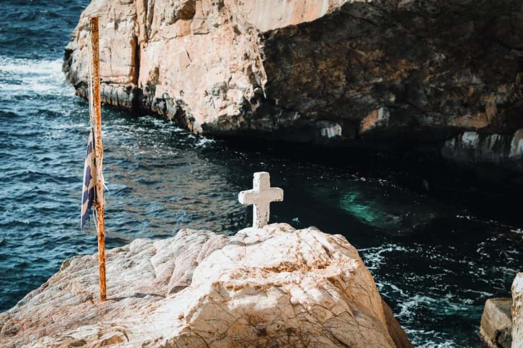 A solitary white cross and a weathered Greek flag stand on a rocky cliff overlooking the dark blue waters off Syros Island, Greece. The rugged coastline and crashing waves create a dramatic, peaceful scene.