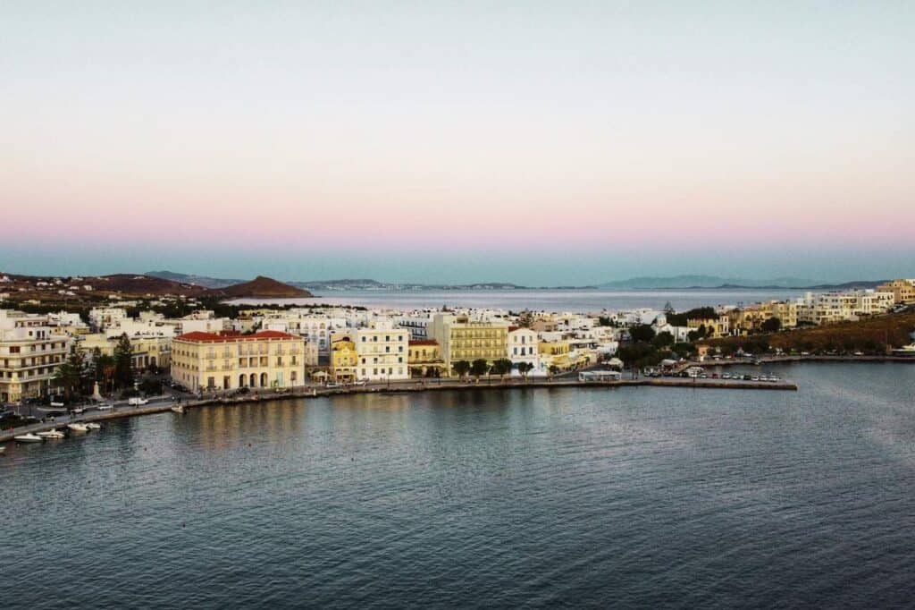 Evening view of Tinos Island, Greece, with pastel hues in the sky reflecting over a calm harbor lined with elegant buildings. The waterfront promenade curves around the bay, capturing the peaceful ambiance of this coastal town as dusk settles.