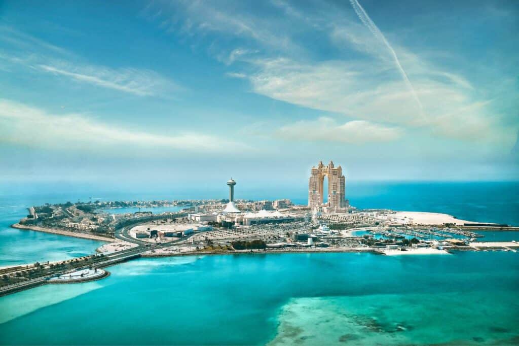 panoramic view of Abu Dhabi, showcasing the turquoise waters of the Persian Gulf, the iconic Marina Mall Tower, and the luxurious Emirates Palace Hotel under a soft blue sky with wispy clouds