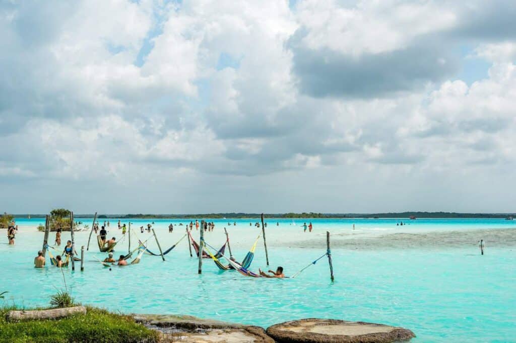 A serene lagoon scene with people relaxing in colorful hammocks suspended over shallow, turquoise water. Others wade and swim in the calm, crystal-clear lagoon, under a sky filled with soft, fluffy clouds. The tranquil setting suggests a laid-back, tropical vibe in Bacalar, Mexico