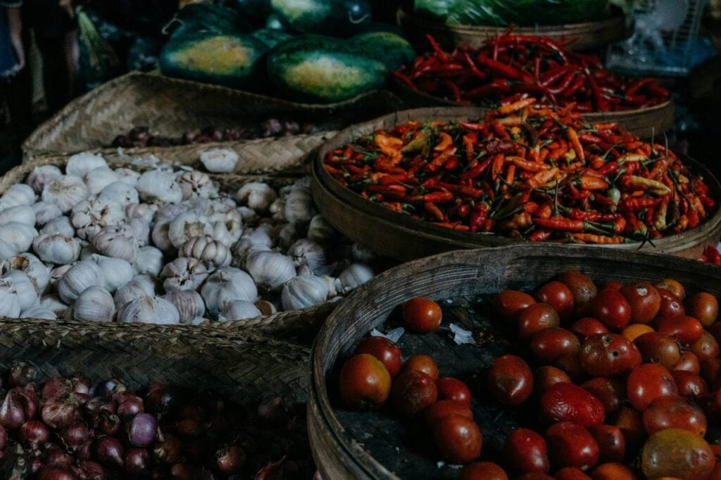 Close-up of various fresh produce displayed at a Bali food market, including baskets of garlic, red chili peppers, tomatoes, and shallots. The vibrant colors and rustic presentation reflect the lively atmosphere of traditional Indonesian markets.
