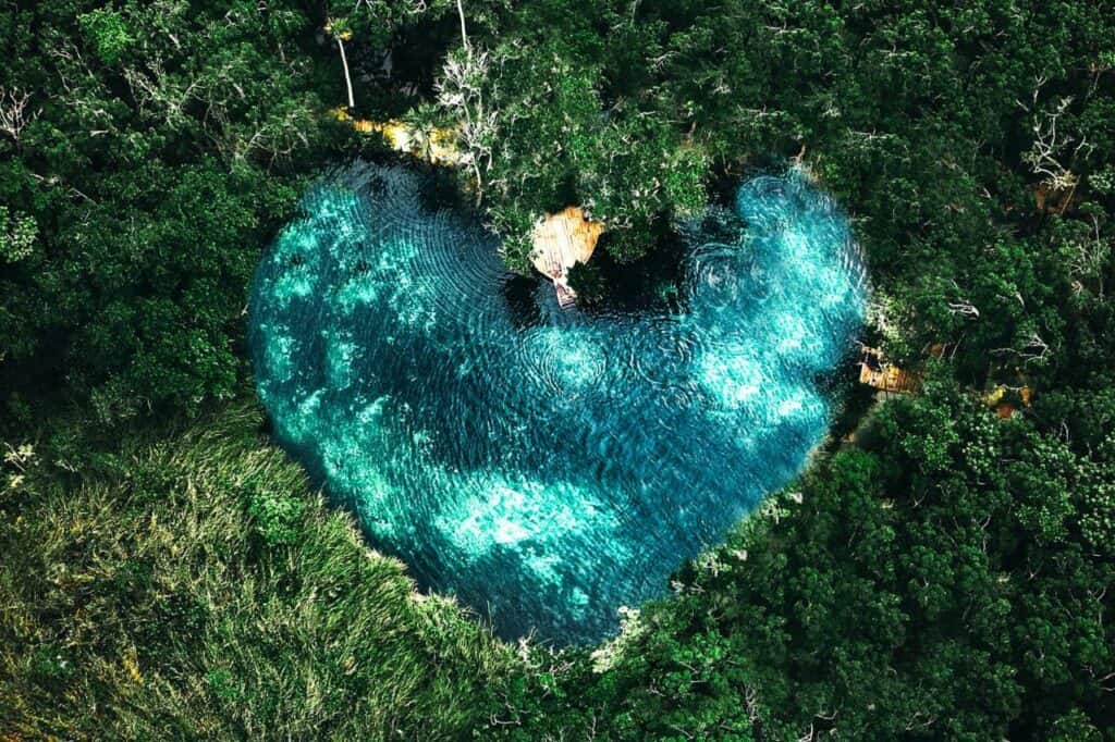 An aerial view of a heart-shaped cenote corazon del paraiso tulum, its crystal-clear turquoise waters surrounded by lush green jungle. The natural pool shimmers under the sunlight, creating a serene and picturesque oasis hidden within the dense foliage.