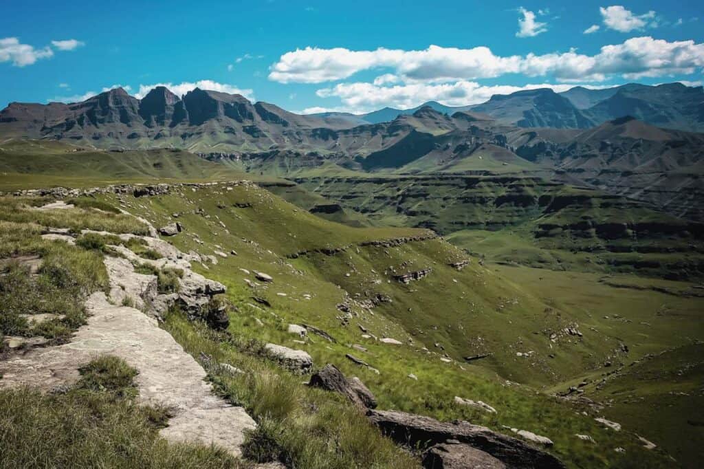 the majestic Drakensberg Mountains in South Africa. The landscape features rugged cliffs and rocky outcrops leading into vast green valleys, with rolling hills stretching toward towering mountain peaks in the background. The clear blue sky with scattered clouds adds to the dramatic beauty of this natural scene.