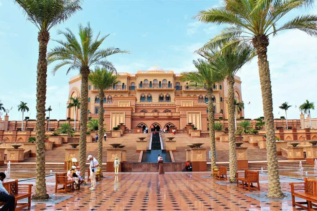 Sunset view at the St. Regis Hotel in Abu Dhabi, with the silhouette of palm trees and sun loungers around a reflective pool capturing the vibrant orange and pink hues of the sky, creating a tranquil and luxurious atmosphere.