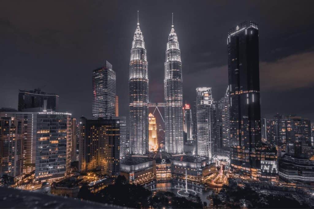 the iconic Petronas Towers in Kuala Lumpur, Malaysia, illuminated against the night sky. The twin towers stand tall, their bright lights highlighting the intricate design, surrounded by other modern skyscrapers. The cityscape below is dotted with lights, adding to the vibrant, bustling atmosphere of Kuala Lumpur's skyline.