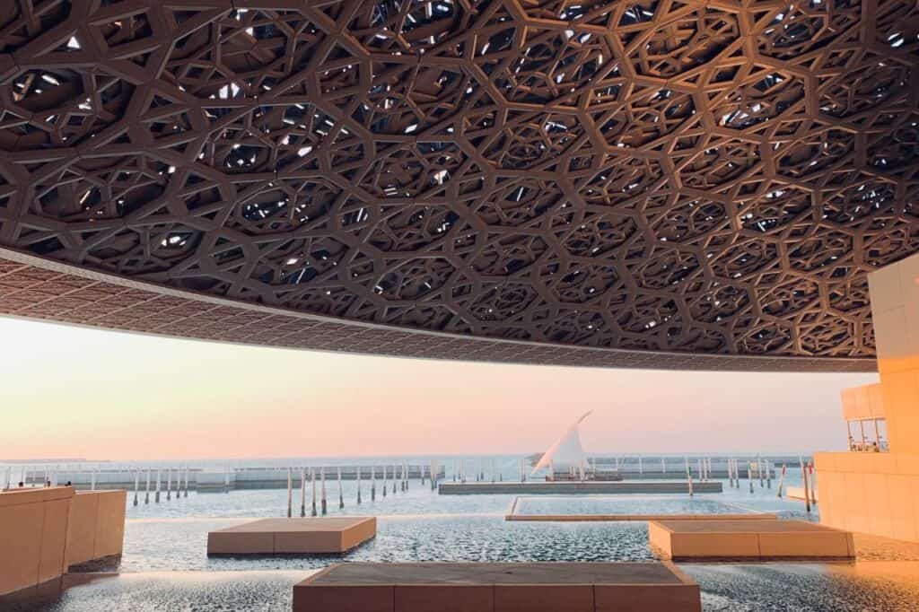 Underneath the intricate geometric dome of the Louvre Abu Dhabi, the view at sunset captures the tranquil sea and the silhouette of the UAE Presidential Palace's flagpole in the distance, all bathed in a warm, ambient light