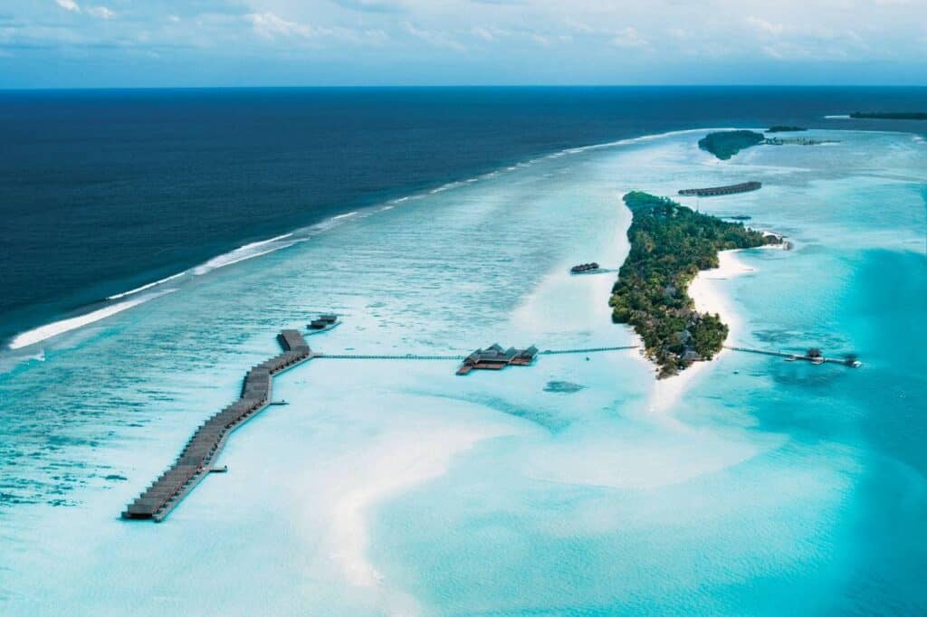 Aerial view of LUX South Ari Atoll Maldives, showcasing overwater villas arranged in a zigzag pattern extending from the island into the shallow, clear turquoise lagoon. The lush, green island is surrounded by pristine white sand beaches, with deeper blue ocean waters in the background.