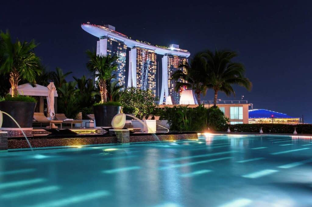 This nighttime image captures the luxurious Marina Bay Sands in Singapore, viewed from a nearby rooftop pool. The iconic Marina Bay Sands building, with its boat-like structure atop the towers, is illuminated against the night sky. The pool in the foreground is surrounded by palm trees and elegant lounge chairs, with soft lighting reflecting off the water