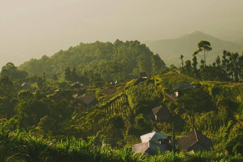 Sunrise view over Mount Batur in Bali, Indonesia, with small traditional houses scattered across the lush green hillsides. The misty mountains and vibrant greenery create a peaceful and serene landscape, popular for hiking and one of the top things to do in Bali on honeymoon.