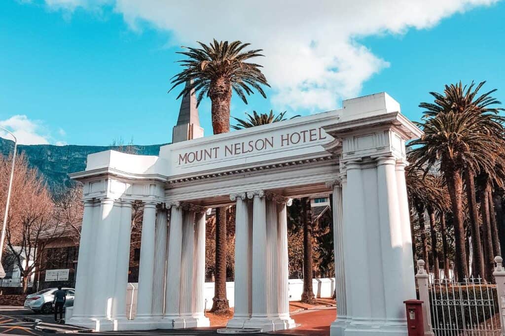 the exterior of the Mount Nelson Hotel in Cape Town, South Africa. The hotel features a grand, colonial-style building painted in pale pink with white trim. Lush green gardens and tall palm trees frame the entrance, with the majestic Table Mountain visible in the background under a clear blue sky.