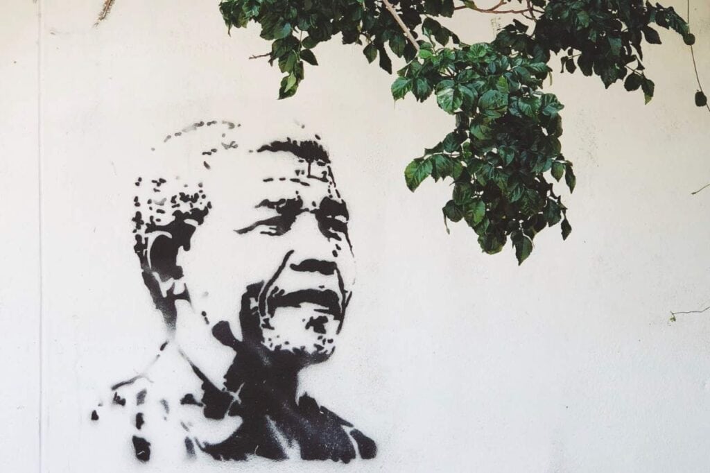 Vibrant street art of Nelson Mandela on a building wall in South Africa. The mural shows a colorful portrait of Mandela smiling, with bold, expressive brushstrokes and a rainbow-like palette. The background is a mix of geometric patterns and bright colors, adding energy and liveliness to the scene.