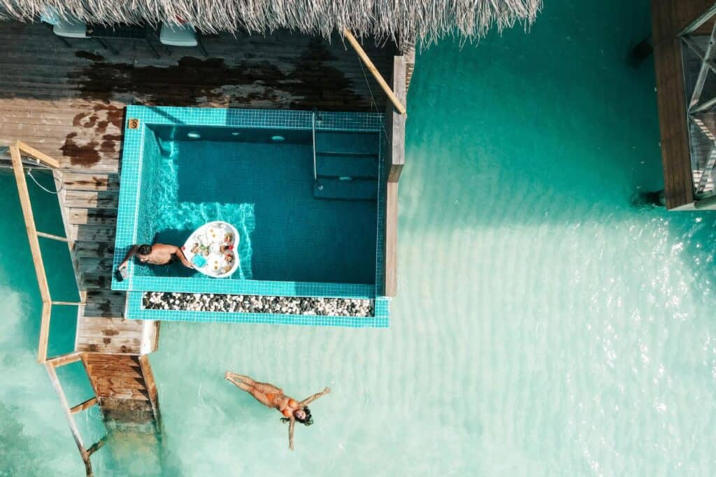 Aerial view of an overwater villa with a private pool; one person enjoys a floating breakfast tray while another relaxes in the shallow lagoon, with vibrant turquoise water surrounding the setup.