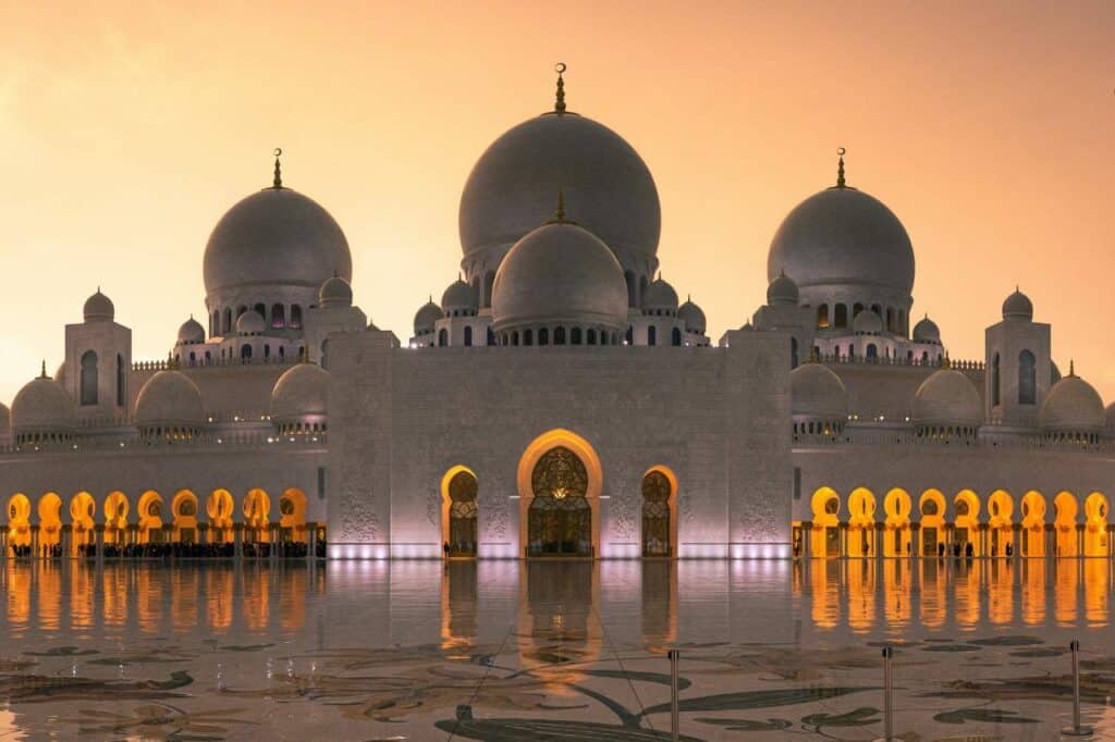 The Sheikh Zayed Grand Mosque in Abu Dhabi captured at twilight, with its white domes and minarets reflecting the warm glow of the setting sun against a serene sky, and its archways casting golden reflections on the polished courtyard