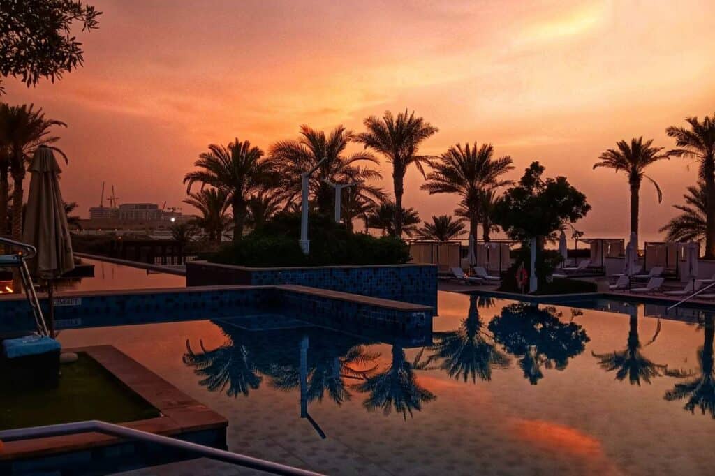 Sunset view at the St. Regis Hotel in Abu Dhabi, with the silhouette of palm trees and sun loungers around a reflective pool capturing the vibrant orange and pink hues of the sky, creating a tranquil and luxurious Abu Dhabi honeymoon atmosphere.