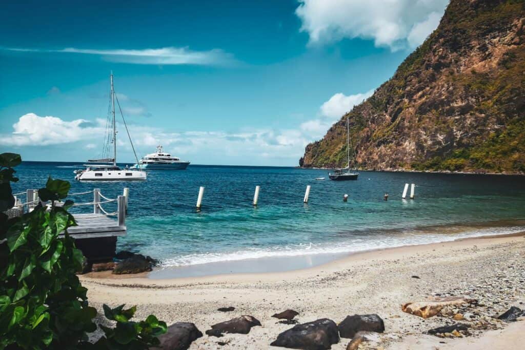 serene view of Sugar Beach in St. Lucia, featuring a sandy shore with scattered rocks, lush green plants near a wooden pier, and two sailboats anchored in the turquoise water. The picturesque scene is framed by a rocky, green-covered cliff under a partly cloudy blue sky.