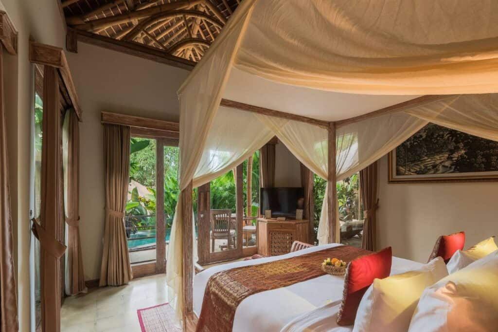 Elegant bedroom in Udaya Resort, Bali, featuring a canopy bed draped with light fabric, natural wooden accents, and large windows opening to a private patio with a view of lush greenery and a private pool.