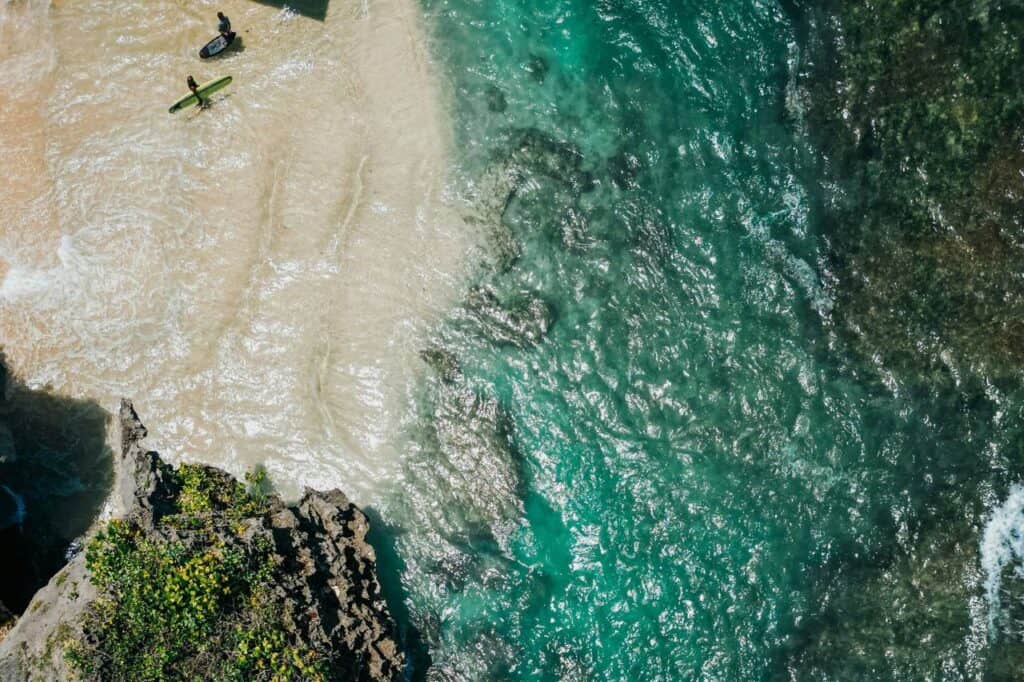 aerial view of Uluwatu Temple in Bali, Indonesia, overlooking the vast blue ocean, with dramatic waves crashing against the rocks below and two surfers near the shore