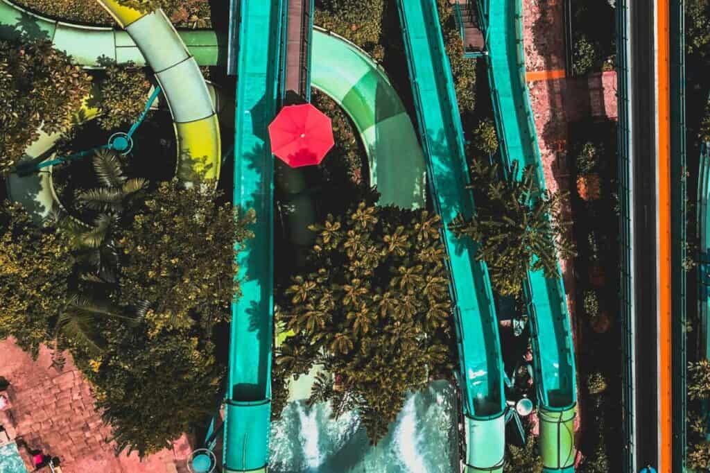 Top-down view of colorful water slides winding through lush greenery at a waterpark, with a bright red umbrella standing out against the green foliage. The image captures the vibrant and fun atmosphere of the park